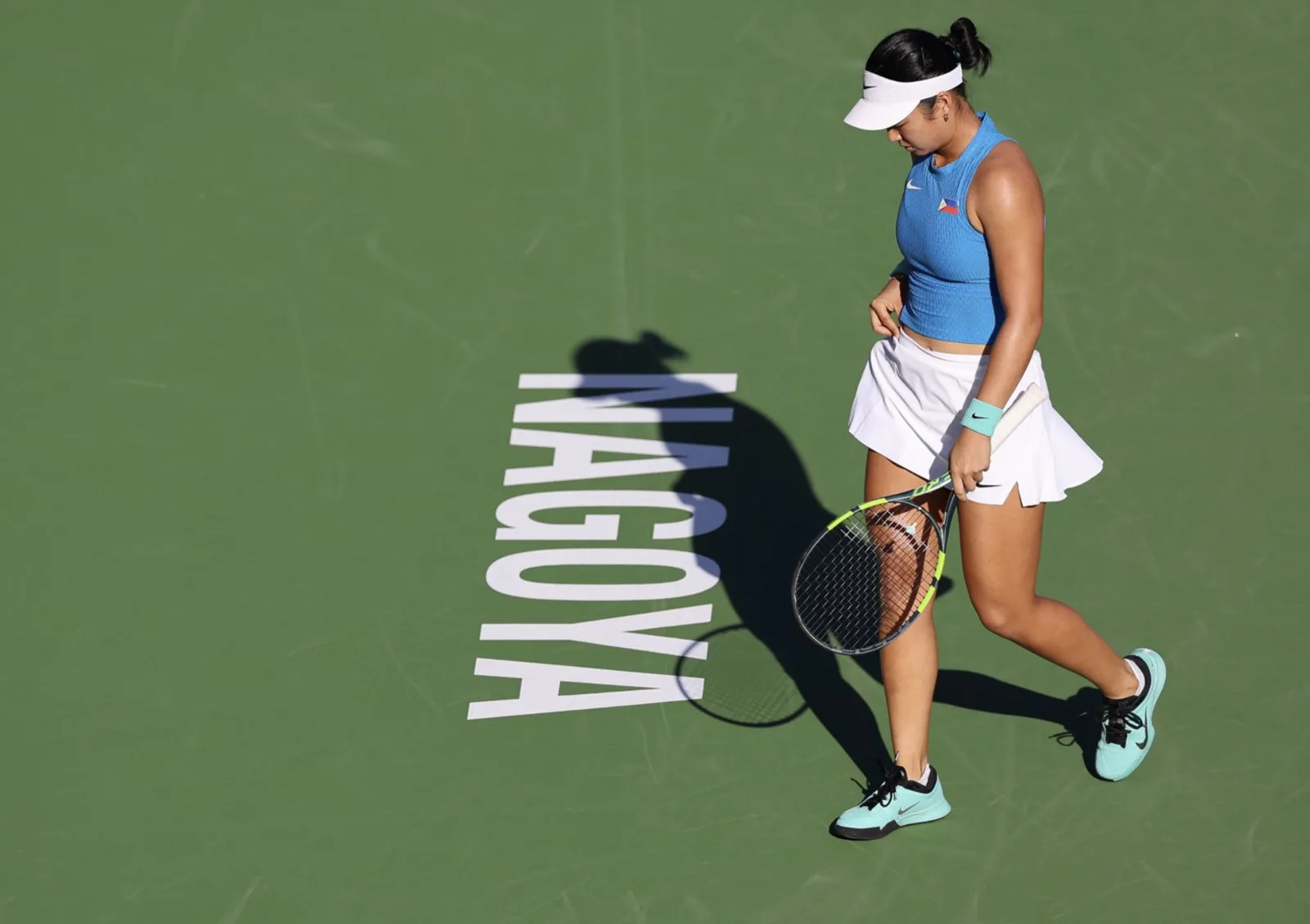 Alex Eala in a blue Philippines top and white visor walks across a green hard court in Nagoya, head down, racket in hand, her shadow falling across the word NAGOYA painted on the court.