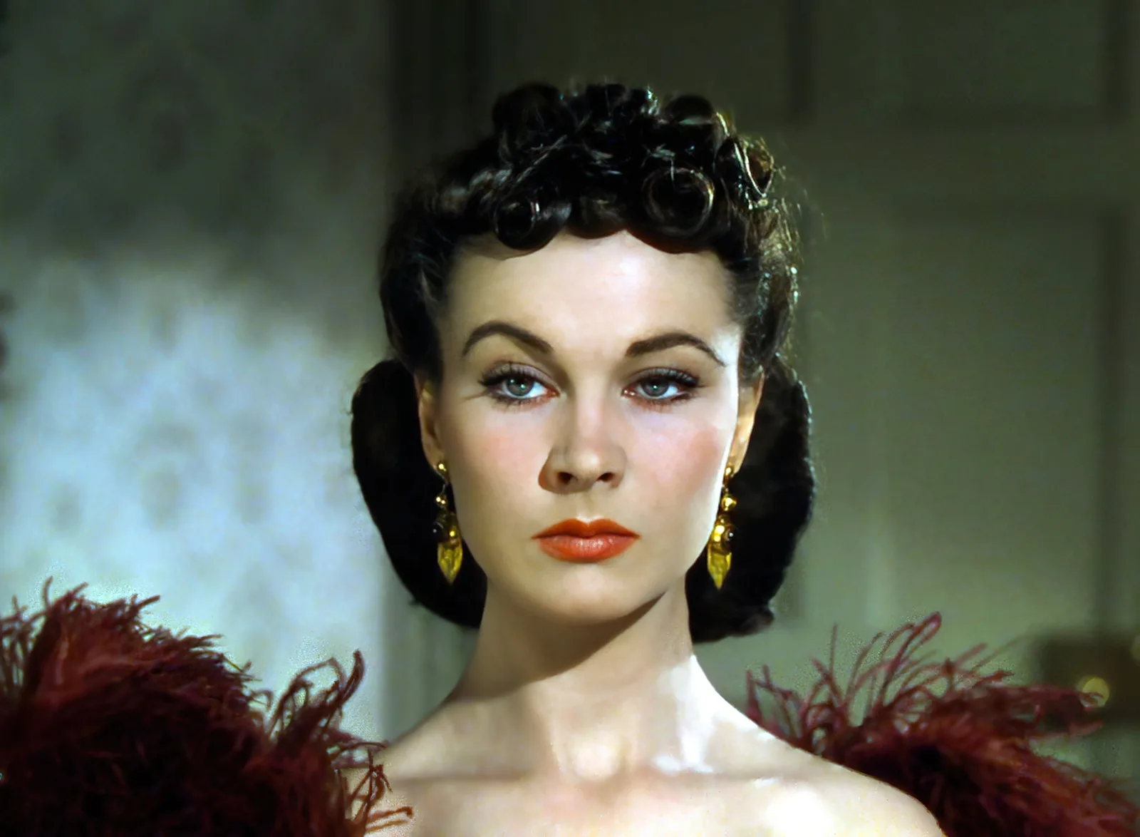 Vivien Leigh as Scarlett O'Hara in close-up, dark curled hair, gold drop earrings and a deep red feather wrap, in a restored color frame from the 1939 Gone with the Wind trailer.