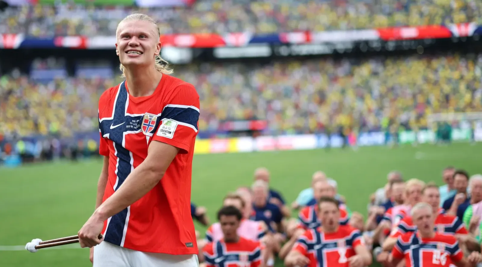 Erling Haaland smiling and holding a drum mallet as Norway's players row in Viking longboat formation behind him.